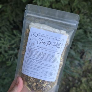 May include: A clear plastic bag containing a herbal tea blend. The label reads "Soothe & Restore Clear the Path Bitter Tea Blend." The bag contains dried herbs and roots. The net weight is 1.7 oz (50g).