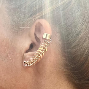 Puede incluir: Un pendiente de aro dorado con un diseño de cadena. El aro se lleva en la parte superior de la oreja y la cadena cuelga.