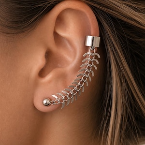 Puede incluir: Un ear cuff plateado con una cadena de eslabones en forma de hoja y un pequeño pendiente de bola. El cuff se envuelve alrededor de la oreja, creando un diseño de joyería único y moderno. La joya es de metal brillante.