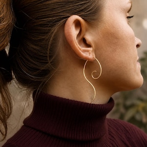 May include: A gold-toned earring featuring a long, slender, S-shaped wire design with a textured surface. The earring curves upward into a spiral at the top and extends downward in a graceful, elongated wave, hanging from the earlobe.