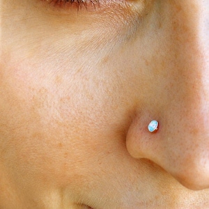 Può includere: Un piccolo piercing al naso rotondo con una pietra di opale liscia e iridescente incastonata in una base metallica. Il gioiello è posizionato sul lato della narice, mostrando un sottile gioco di luce sulla superficie della pietra con toni blu.