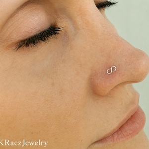 May include: Close-up of a person's nose with a small, silver infinity symbol nose stud. The jewelry is delicate and sits on the nostril. The brand name "KRacz Jewelry" is visible at the bottom of the image.