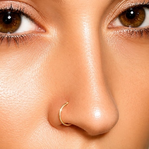 May include: Close-up of a person's face, focusing on the nose and eyes. A gold-colored nose ring is visible on the nostril. The skin tone is light brown, and the eyes are brown with long eyelashes.