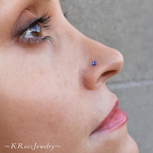 May include: Close-up of a person's face with a blue nose stud. The stud is a small, round, and shiny gemstone. The skin is fair with a slight pink hue on the lips. The text "~KRaczJewelry~" is at the bottom.