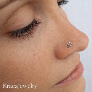 May include: Close-up of a person's face with a silver nose stud. The nose stud has a unique design, featuring three connected loops. The brand name "KraczJewelry" is visible at the bottom of the image.