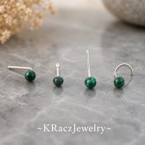 May include: Four nose studs with round, dark green malachite stones and silver-toned metal posts. The studs feature various post shapes, including straight, L-shaped, and curved designs. The text ~KRaczJewelry~ is printed at the bottom of the image.