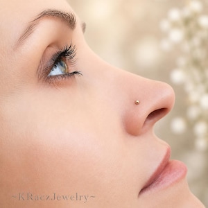 May include: A small, round, gold-toned nose stud featuring a smooth, polished spherical head. The jewelry is positioned on the side of the nostril. The text KRaczJewelry is visible in the bottom left corner of the image.