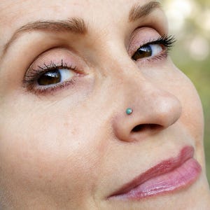 May include: A small, round nose stud featuring a smooth, teal-colored stone set in a metallic base. The jewelry is positioned on the side of the nostril, displaying a simple, circular design with a polished finish and a vibrant, solid color.