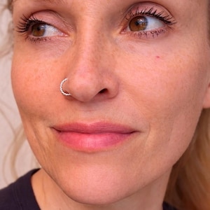 May include: Close-up of a person's face with a silver nose ring. The nose ring has a twisted rope design. The person has light skin, brown eyes, and is looking to the side.