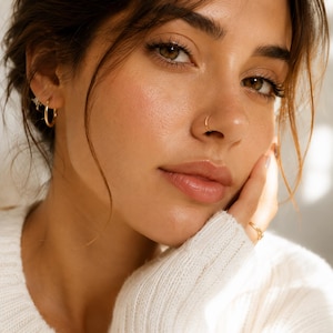 May include: A set of gold jewelry consisting of a small hoop earring in the earlobe, a thin gold nose ring, and a simple gold band ring worn on a finger. The jewelry features a polished metallic finish and a slender, circular design.