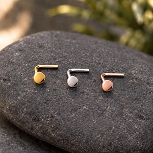 May include: Three L-shaped nose studs with flat circular heads, arranged in a row. The first stud is gold-toned, the second is silver-toned, and the third is rose gold-toned. Each piece features a smooth, polished finish and a bent post design.