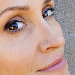 May include: Close-up of a person's face, focusing on a small, round, silver nose stud. The skin is fair with visible pores. The person's eyes are brown, with dark eyeliner and mascara. The background is a plain, gray wall.