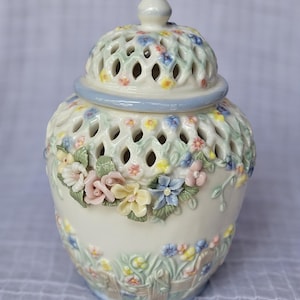 May include: A white ceramic jar with a lid, adorned with colorful floral designs. The jar features a lattice pattern and a light blue trim. The lid has a small knob on top. The jar is decorated with pink, yellow, and blue flowers.