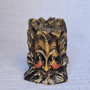 May include: A detailed, carved candle holder in the shape of a bearded face. The face has a yellow nose and cheeks, with dark eyes and a long, flowing beard. The candle holder is primarily brown and gold, with intricate details.