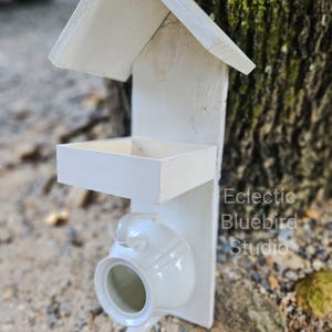 May include: A white wooden bird feeder with a pitched roof, a feeding tray, and a repurposed white teapot. The bird feeder is mounted on a wooden backing and features the text "Eclectic Bluebird Studio".