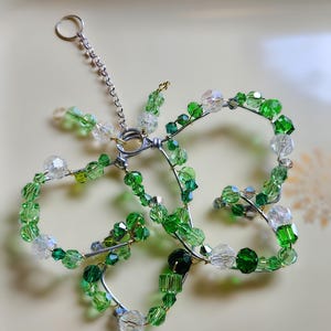 May include: A handcrafted butterfly ornament made of silver wire and green and clear glass beads. The ornament has a small chain and ring for hanging. The beads are various shapes and sizes, creating a sparkling effect.