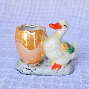 Pode incluir: Estatueta de pato de cerâmica com um recipiente em forma de ovo. O pato tem corpo branco, bico e pés laranja e uma asa verde e laranja. O recipiente para ovos é laranja com um acabamento iridescente dourado. A base é cinza.