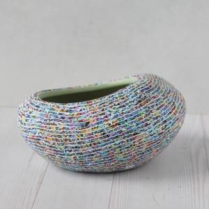 May include: A decorative, oval-shaped ceramic vase with a textured, multi-colored exterior. The vase features a light green interior and is set on a white wooden surface. The exterior design incorporates various colors, creating a unique, artistic appearance.