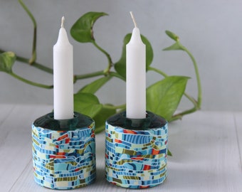 May include: Two white candles in teal blue and green patterned ceramic candle holders. The holders have a geometric design with orange, yellow, and blue accents.