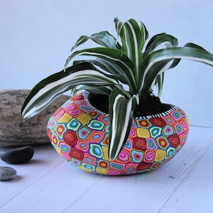 Pode incluir: Um vaso colorido com um padrão geométrico e uma planta verde com listras brancas. O vaso é feito de argila polimérica e tem um design único feito à mão.