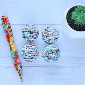 May include: A vibrant pen with a patterned design in red, yellow, and blue, alongside four decorative round objects with a similar design. A small succulent plant in a white pot is also in the image.