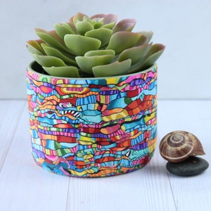 May include: A colorful, abstract patterned ceramic planter with a green succulent plant inside. The planter is sitting on a white wooden surface.