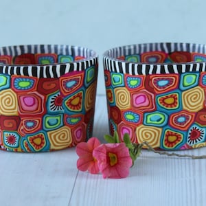 Può includere: Due vasi decorativi, di forma quadrata, con un motivo astratto e colorato. I vasi presentano un bordo a righe bianche e nere. Un rametto di fiori rosa è posto davanti ai vasi.