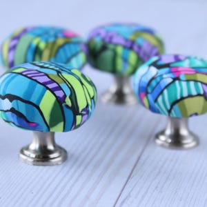 May include: Four colourful fabric-covered cabinet knobs with silver bases. The knobs feature a geometric pattern in shades of blue, green, purple, and pink.