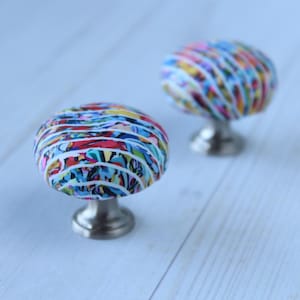 May include: Two decorative cabinet knobs with a colorful, abstract design. The knobs have a rounded shape with a base made of silver-toned metal. The colorful design features a mix of red, blue, yellow, and white stripes.