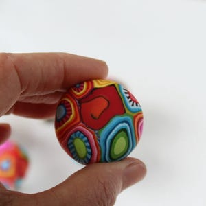 May include: A colourful, round, patterned clay bead with red, blue, green, yellow, and white swirls. The bead is being held in a hand.