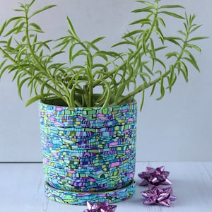 May include: A green succulent plant in a blue and green patterned ceramic pot with a matching saucer. The pot has a wavy, abstract design with pink, purple, and green accents. There are three pink bows on the white surface.