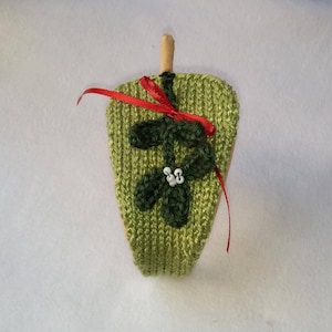 May include: A green knitted ornament shaped like a mistletoe sprig with white beads for berries and a red ribbon tied around the stem.