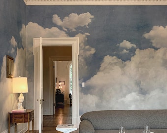 Puede incluir: Una habitación con paredes cubiertas de papel tapiz de cielo azul con nubes blancas. El espacio incluye un sofá gris, una mesa de centro negra con cristalería, una mesa auxiliar de mármol blanco y una mesa de madera con una lámpara blanca.