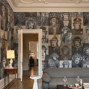 Puede incluir: Una habitación con un sofá gris, una mesa de centro negra y papel pintado con motivos de monos. El papel pintado presenta una cuadrícula de retratos de monos con diferentes atuendos. También se ve una mesa auxiliar de mármol y una mesa auxiliar de madera con una lámpara.