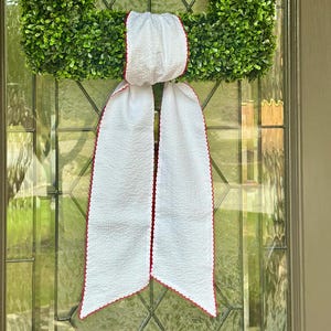 May include: A square, green boxwood wreath hangs on a glass door. A white ribbon with red trim is tied to the wreath. The door has a decorative glass design and is framed by a dark brown door frame.