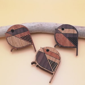 May include: Three wooden bird brooches with geometric designs. Each brooch features light and dark wood tones, with black accent lines. The birds have a circular body and a long, thin tail. The brooches are displayed on a light-coloured background.
