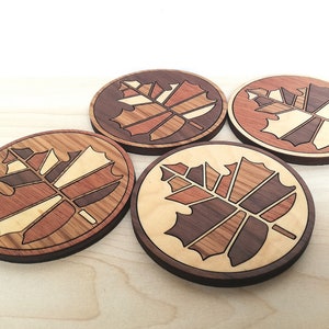 May include: Four wooden coasters with a maple leaf design. The coasters are made of different types of wood, creating a unique and intricate pattern.