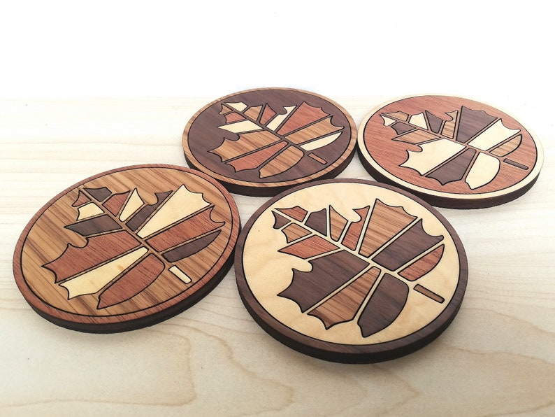 May include: Four wooden coasters with a maple leaf design. The coasters are made of different types of wood, creating a unique and intricate pattern.