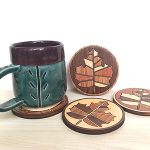 May include: Three wooden coasters with a maple leaf design in shades of brown, red, and yellow. The coasters are round and have a dark brown border. A green and brown ceramic mug is sitting on one of the coasters.