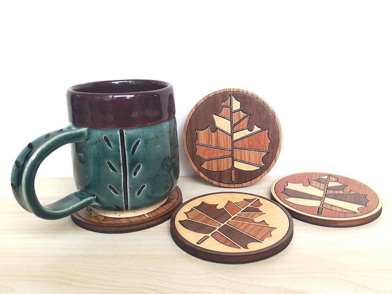 May include: Three wooden coasters with a maple leaf design in shades of brown, red, and yellow. The coasters are round and have a dark brown border. A green and brown ceramic mug is sitting on one of the coasters.