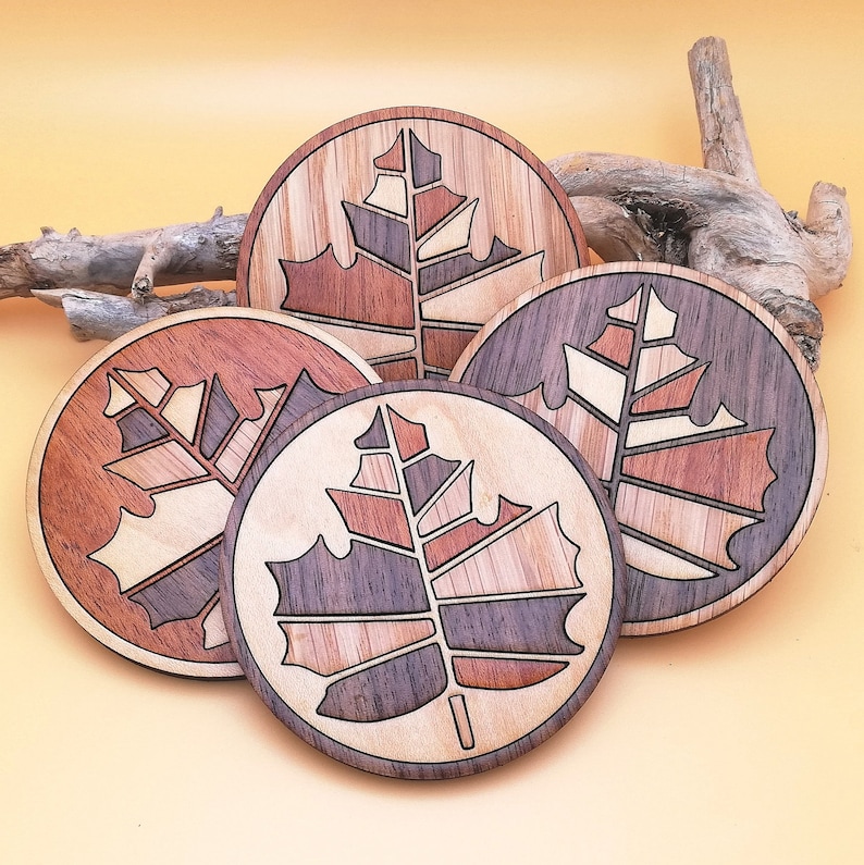 May include: Four round wooden coasters with a laser-cut design of a leaf in shades of brown, beige, and black.