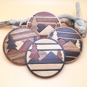 May include: Set of four wooden coasters featuring a mountain range, trees, and a sun. Each coaster has a different colour scheme, with shades of brown, black, and white.