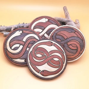 Peut inclure: Quatre sous-verres en bois avec un motif de deux serpents entrelacés. Les serpents sont sculptés dans un bois clair et foncé, créant un motif contrasté. Les sous-verres sont ronds et ont une finition lisse.