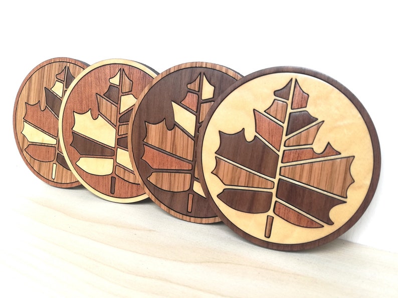 May include: Four round wooden coasters with a maple leaf design. Each coaster is made of different types of wood, including walnut, maple, and cherry. The coasters are arranged in a row, with the darkest coaster on the right and the lightest coaster on the left.