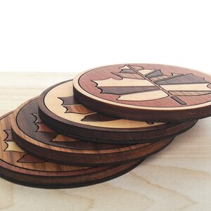 May include: Set of four wooden coasters featuring a maple leaf design. The coasters are made of different types of wood, creating a unique and colorful pattern.