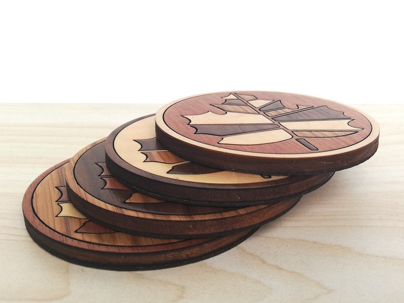 May include: Set of four wooden coasters featuring a maple leaf design. The coasters are made of different types of wood, creating a unique and colorful pattern.