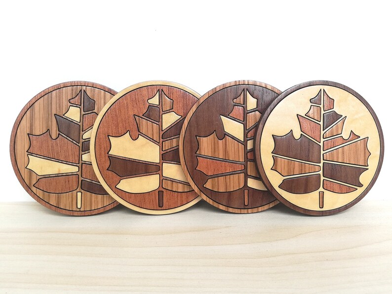May include: Four wooden coasters with a maple leaf design. Each coaster features a different wood grain pattern, creating a unique and natural look.
