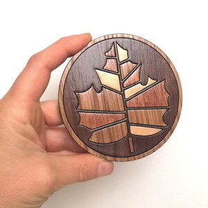 May include: A round wooden coaster with a carved maple leaf design. The leaf is made of different types of wood, creating a multi-colored effect.