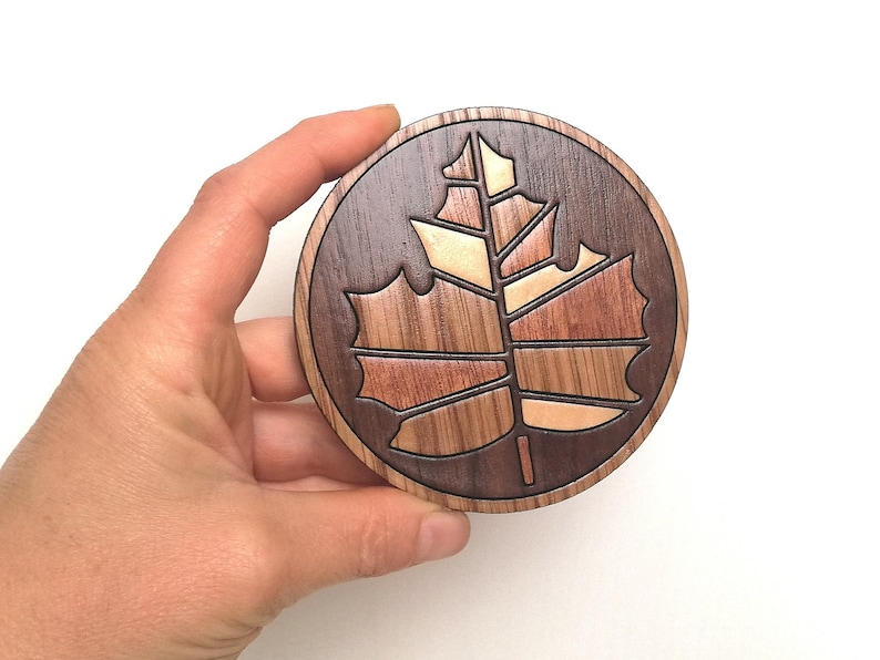 May include: A round wooden coaster with a carved maple leaf design. The leaf is made of different types of wood, creating a multi-colored effect.