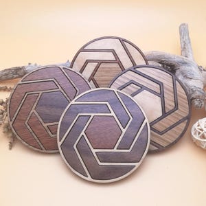 May include: Five wooden coasters with geometric patterns. Each coaster is circular, featuring a central hexagon and surrounding angular shapes, made from different shades of brown and tan wood. They are arranged on a light beige surface.